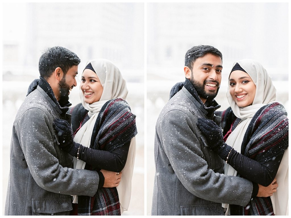 A couple embraces while looking at each other and then at the camera. They are covered with snow flurries. 