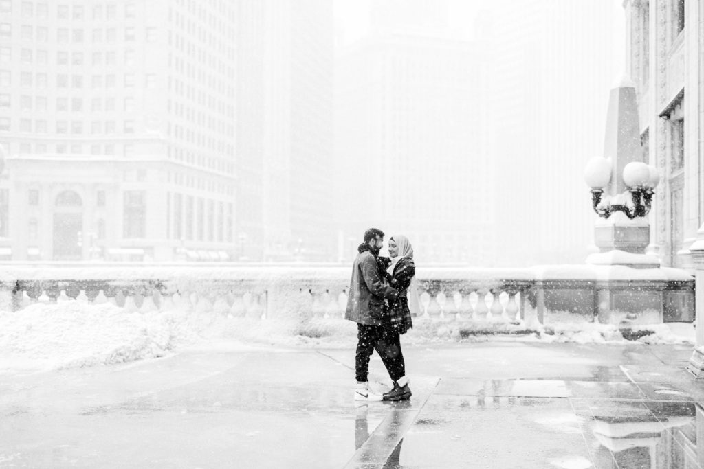 A hugging couple gaze at each other with tall snow filled buildings in the background. 