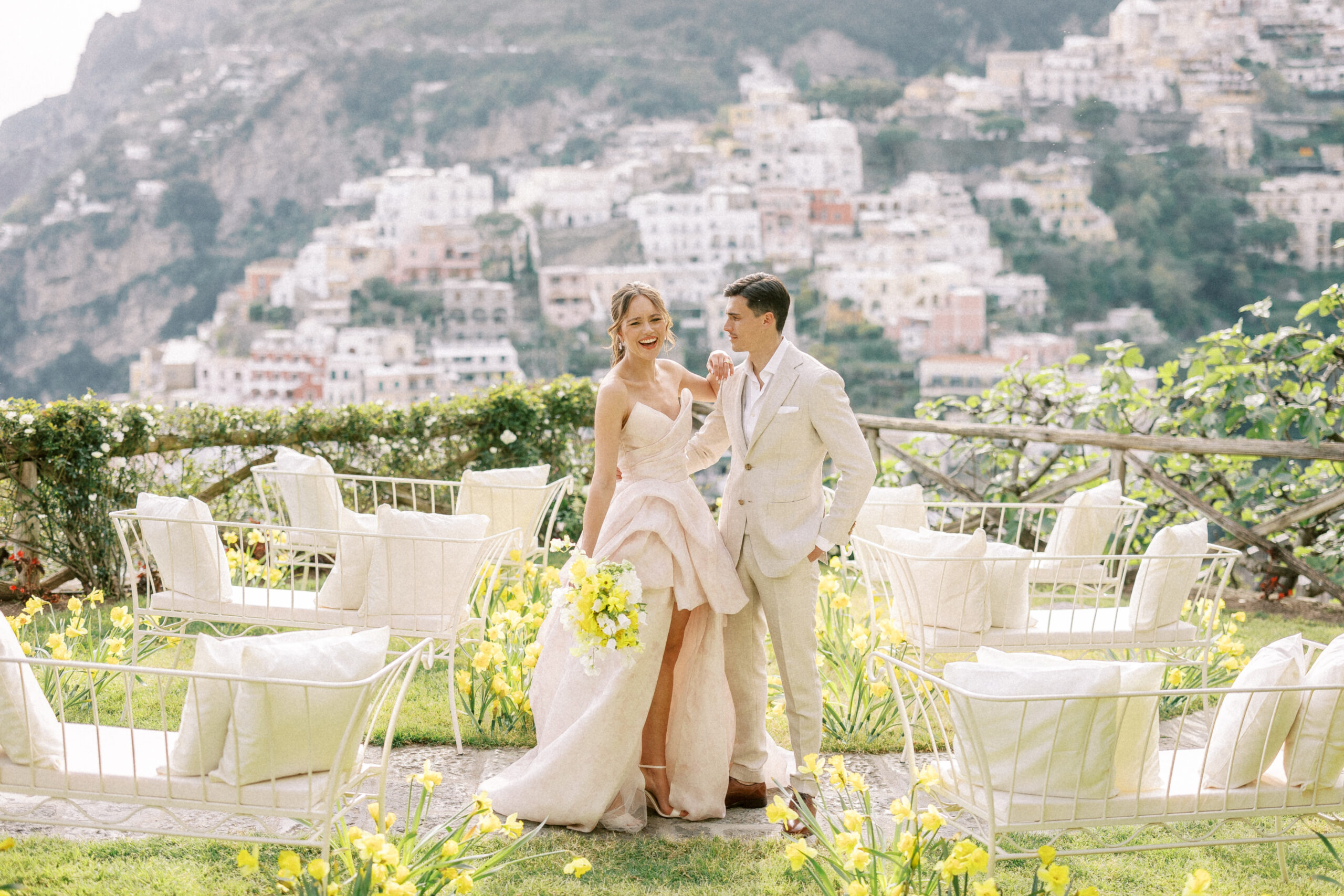 A groom looks at his bride while standing between their ceremony setup of white couches and a runway of daffodils. The background is slightly blurred but showcases villas on an Italian hillside.