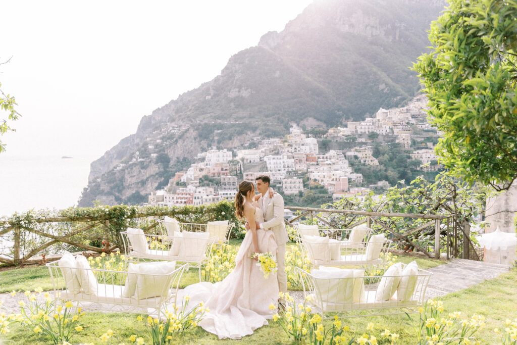 A fashionable couple embraces at a private Positano hillside villa. An array of colorful Italian homes are in the distant background. 