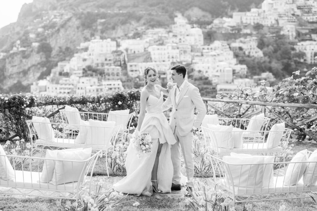 The bride laughs toward the camera while holding the bouquet. The groom embraces the bride while his left hand is in the pant pocket of his tailored Italian suit. Positano homes are blurred in the background. This image is edited in black and white. 