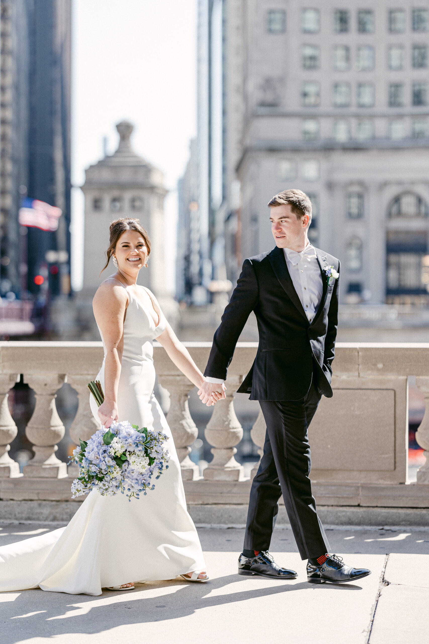 A wedding couple walks hand-in-hand down a near Chicago's riverwalk. They are both smiling and laughing, and they look very happy together. The sun is shining, and the bride is smiling at the camera while holding her bouquet. Her new husband glances back at her while smiling. It is a beautiful day, and the couple is enjoying each other's company.